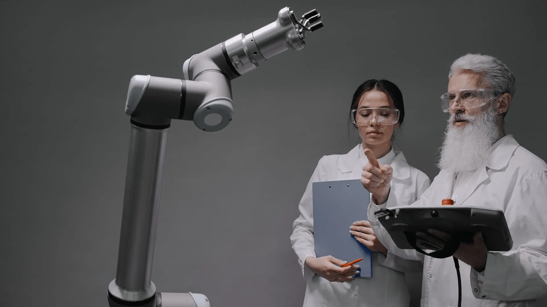 A man with beard and a girl in scientist apron looking at a robotic arm